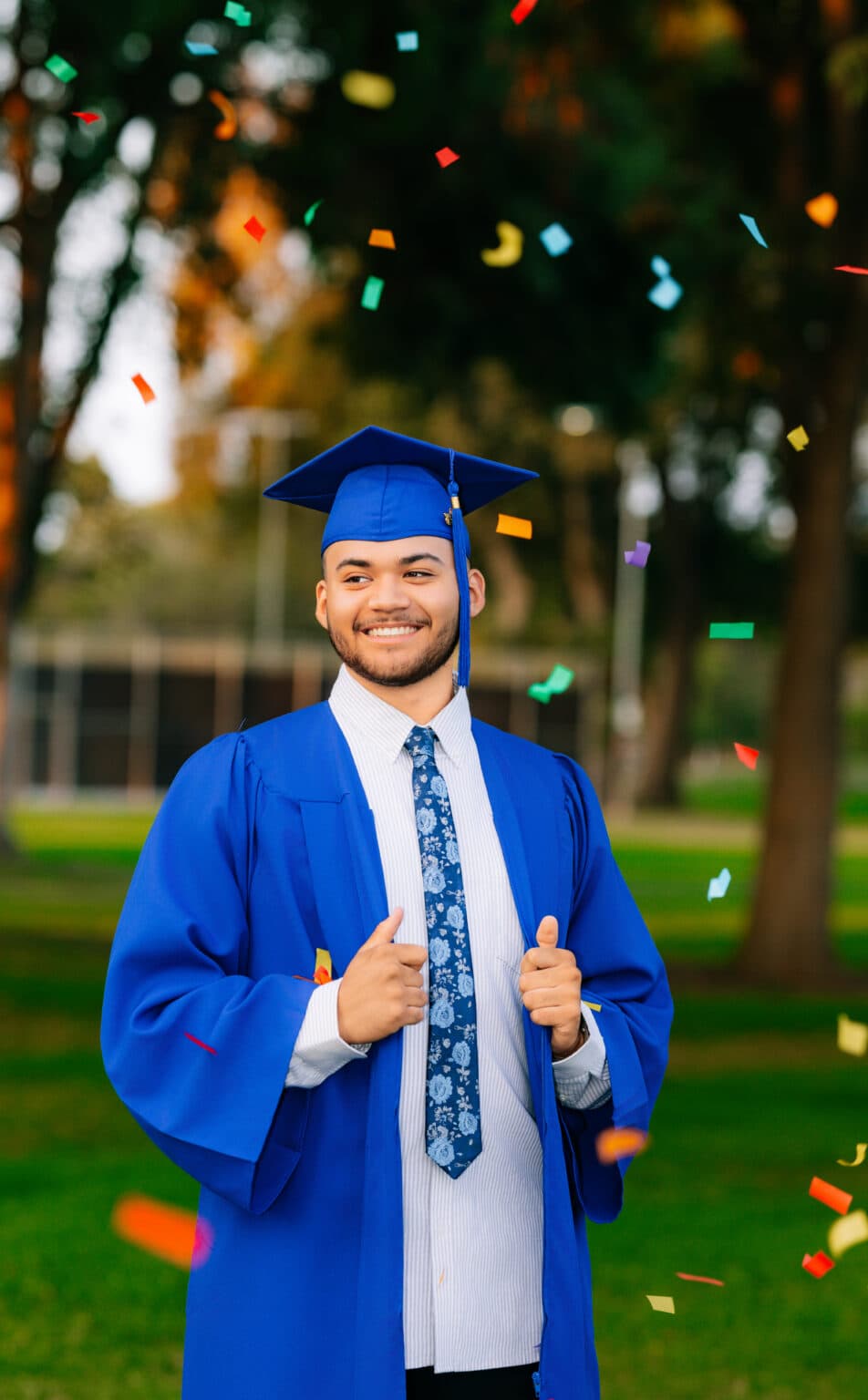 Smiling male graduate in a blue cap and gown celebrating with falling confetti during a Southern California senior portrait session.